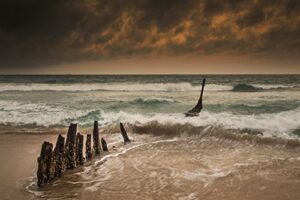 posterazzi wooden posts on a beach with a boat being tossed in the water and waves crashing into the sand under a cloudy sky queensland australia poster print, (19 x 12)